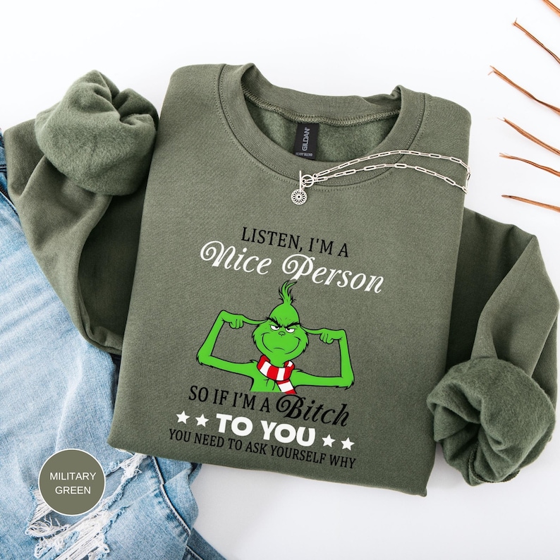 Op de afbeelding: Een olijfgroene sweatshirt met de tekst "LISTEN, I'M A Nice Person" en "SO IF I'M A Bitch TO YOU YOU NEED TO ASK YOURSELF WHY". De afbeelding toont een groen stripfiguur met een rood-witte sjaal.