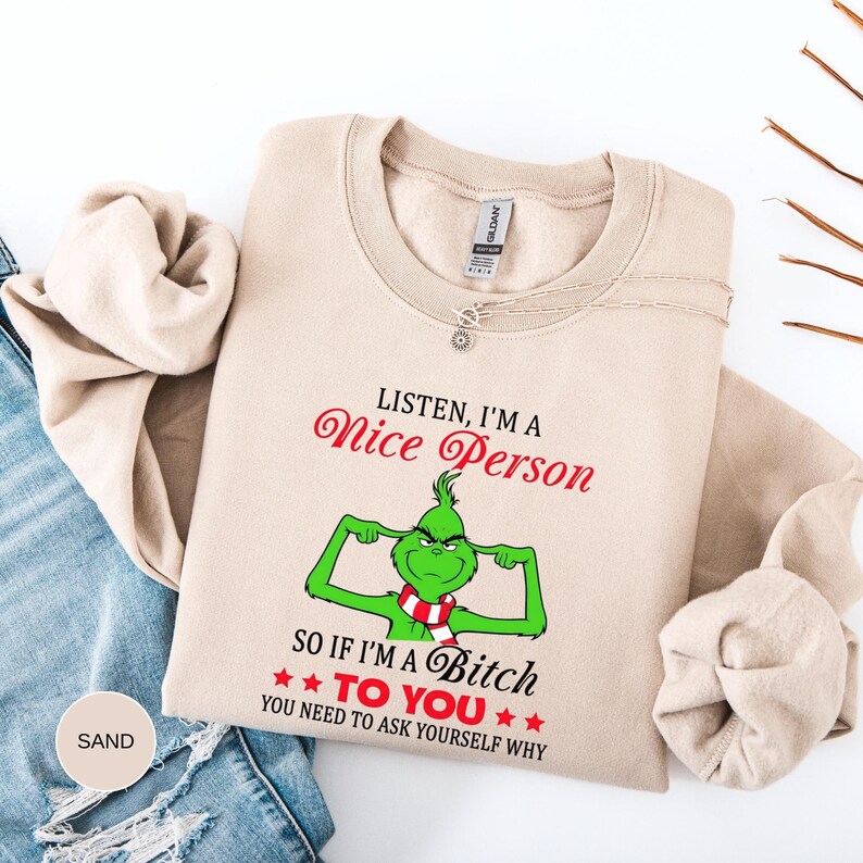 Op de afbeelding: Een zandkleurige sweatshirt met de tekst "LISTEN, I'M A Nice Person" in rode letters. Daaronder staat een groen stripfiguur met een rood-witte sjaal en de tekst "SO IF I'M A Bitch TO YOU YOU NEED TO ASK YOURSELF WHY".