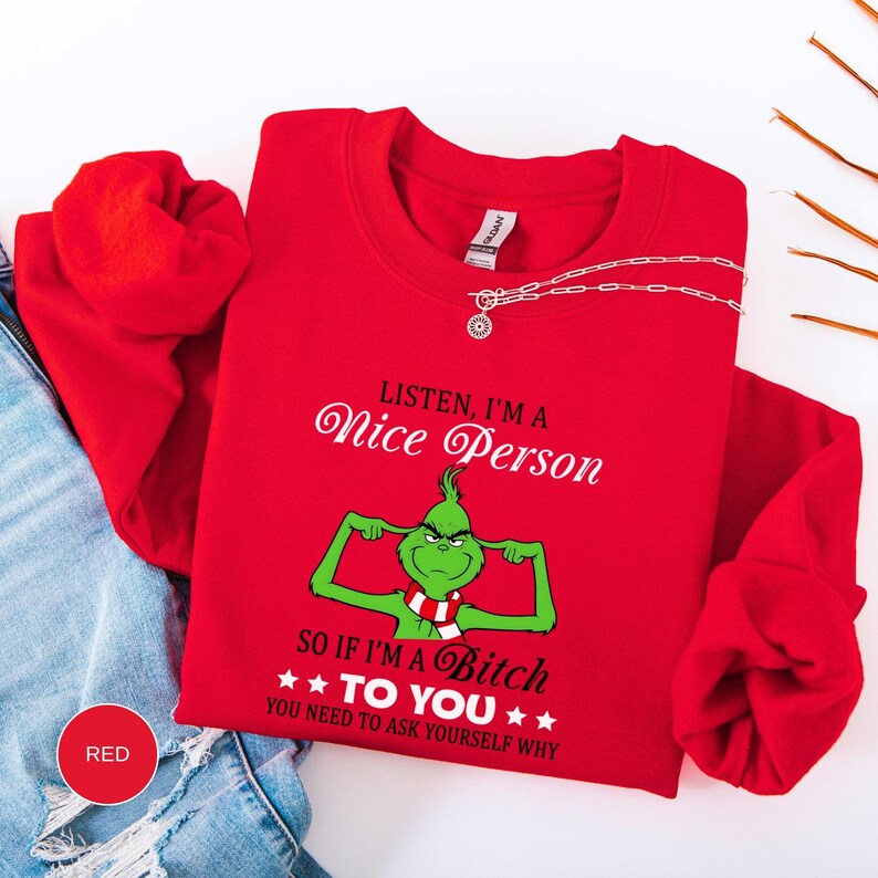 Op de afbeelding: Een rode crewneck sweatshirt met de tekst "Listen, I'm a Nice Person, so if I'm a Bitch to You, You Need to Ask Yourself Why." Een groen stripfiguur met een rood-witte sjaal poseert. Een zilveren ketting ligt op de sweatshirt.