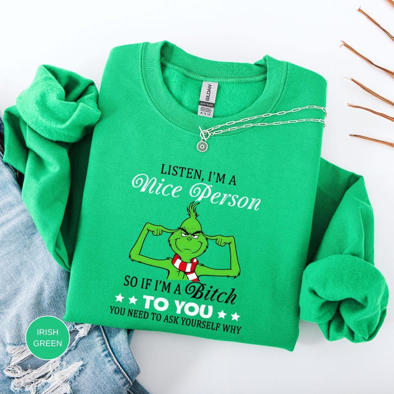 Op de afbeelding: Iers groen sweatshirt met een Grinch-afbeelding en de tekst "Listen, I'm a Nice Person, so if I'm a Bitch to you, you need to ask yourself why." Een zilveren ketting ligt over het sweatshirt.