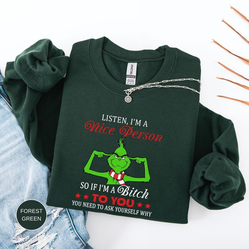 Op de afbeelding: Bostgroen sweatshirt met de tekst "LISTEN, I'M A Nice Person" in rode letters. Daaronder staat een groene stripfiguur die spieren laat zien, met de tekst "SO IF I'M A Bitch TO YOU YOU NEED TO ASK YOURSELF WHY". Een zilveren ketting ligt op het sweatshirt.