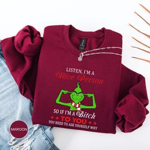 Op de afbeelding: Bordeaux crewneck sweatshirt met de tekst "LISTEN, I'M A Nice Person" en een groen cartoonfiguur. Daaronder staat de tekst "SO IF I'M A Bitch TO YOU YOU NEED TO ASK YOURSELF WHY". Een zilveren ketting ligt op de sweatshirt.