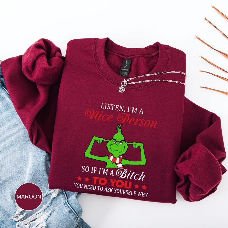 Op de afbeelding: Bordeaux crewneck sweatshirt met de tekst "LISTEN, I'M A Nice Person" en een groen cartoonfiguur. Daaronder staat de tekst "SO IF I'M A Bitch TO YOU YOU NEED TO ASK YOURSELF WHY". Een zilveren ketting ligt op de sweatshirt.