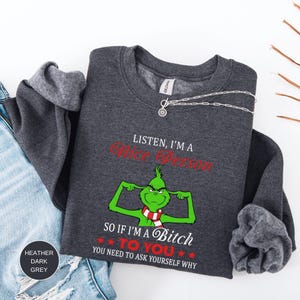 Op de afbeelding: Donkergrijze sweatshirt met de tekst "LISTEN, I'M A Nice Person" in het rood en "SO IF I'M A Bitch TO YOU YOU NEED TO ASK YOURSELF WHY" in het rood. De sweatshirt heeft een groen stripfiguur met een rood-witte sjaal.
