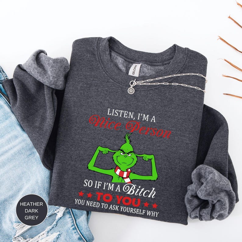 Op de afbeelding: Donkergrijze sweatshirt met de tekst "LISTEN, I'M A Nice Person" in het rood en "SO IF I'M A Bitch TO YOU YOU NEED TO ASK YOURSELF WHY" in het rood. De sweatshirt heeft een groen stripfiguur met een rood-witte sjaal.