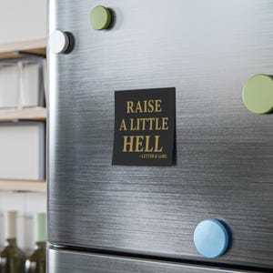 Könnte beinhalten: Schwarzer und goldener Kühlschrankmagnet mit dem Text "Raise a little hell - Letter & Lore".