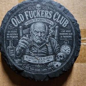 Op de afbeelding: Een ronde, donkergrijze leistenen onderzetter met de tekst "OLD FUCKERS CLUB" en een illustratie van een oudere man die een sigaar rookt en drinkt. Extra tekst bevat "AGED TO PERFECTION" en "RESPECT YOUR ELDERS (OR ELSE)".
