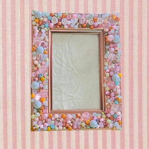 May include: A rectangular photo frame with a rose gold finish, embellished with colorful beads in shades of pink, orange, blue, and clear. The word "BUTTON" is spelled out across the top of the frame with letter beads.