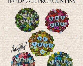 Puede incluir: Broches de pronombres hechos a mano con diseños de cuentas. Los broches presentan los pronombres "He/Him", "She/Her" y "They/Them" escritos con letras blancas sobre un fondo de cuentas de colores. El texto "Handmade Pronoun Pins" está en la parte superior.