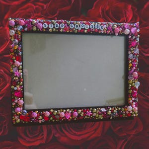 May include: A black rectangular picture frame adorned with pink, red, and gold beads, gems, and pearls. The top of the frame features the words "STAR CROSSED" in white letter beads. The frame is set against a backdrop of red roses.