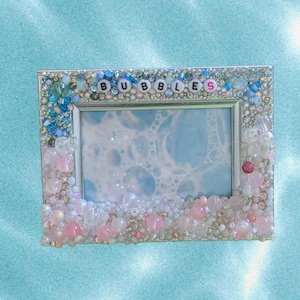 May include: A rectangular photo frame with a beaded border. The frame is decorated with clear, pink, and blue beads. The word "BUBBLES" is spelled out across the top of the frame in letter beads. The photo inside the frame has a blue and white bubble design.