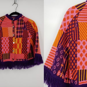 May include: A colorful knitted poncho with a geometric pattern in shades of pink, orange, yellow, and purple. The poncho has a button closure and a fringe trim.