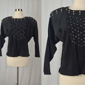 Puede incluir: Top negro de manga larga con adornos. El top presenta un escote redondo y está adornado con pequeños pedrería plateada en un patrón curvo en la parte delantera. Tachuelas doradas están colocadas en los hombros. Las mangas son largas y la tela es suave.