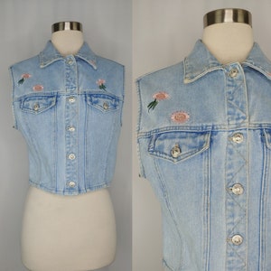 May include: A light blue denim vest with a button-up front and embroidered flowers. The vest has two pockets on the front and a collar.