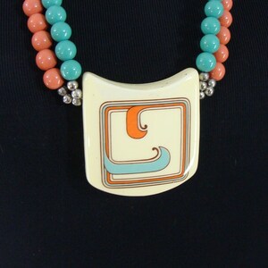 May include: A white ceramic pendant necklace with a geometric design in orange, blue, and grey. The pendant is attached to a double strand of coral and turquoise beads.
