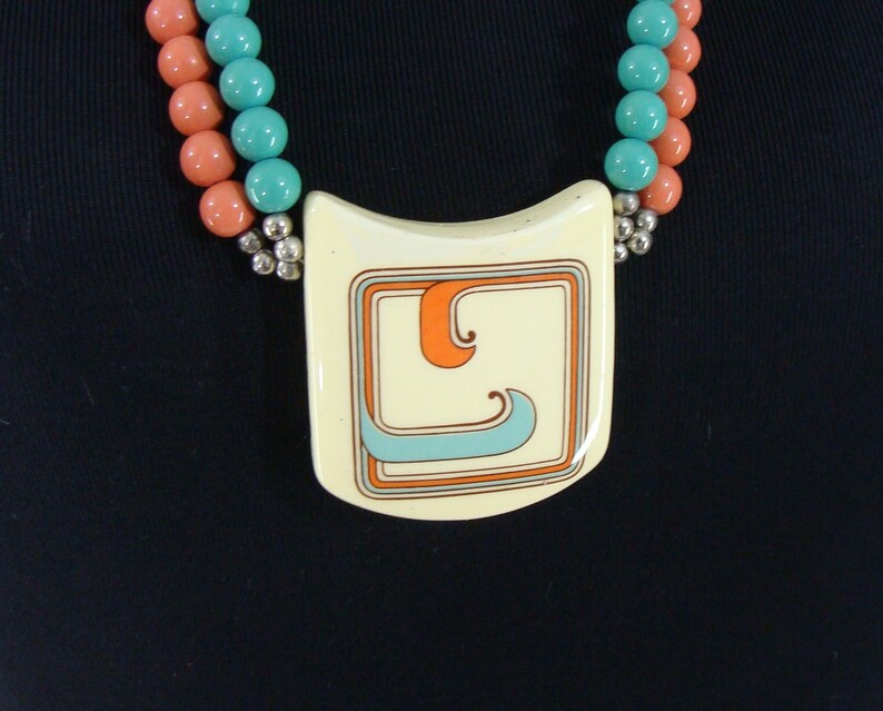 May include: A white ceramic pendant necklace with a geometric design in orange, blue, and grey. The pendant is attached to a double strand of coral and turquoise beads.