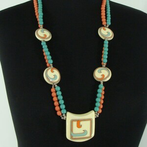 May include: A double-strand necklace with a large, square pendant. The necklace is made of turquoise and coral-coloured beads. The pendant is white with a blue and orange swirl design.