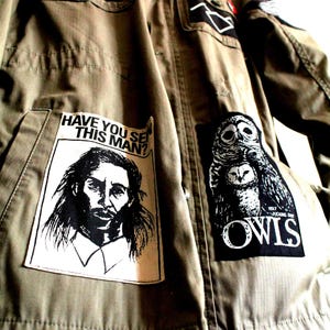 May include: Olive green jacket with multiple patches. Patches include text and illustrations of a man, owls, and other graphics. The jacket has multiple pockets and buttons.