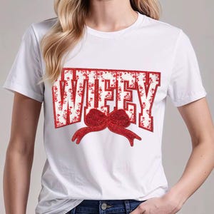 Op de afbeelding: Wit T-shirt met het woord "WIFEY" in rode blokletters met een bloemenpatroon. Een rode strik staat eronder. Het shirt heeft een ronde hals.