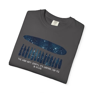May include: Dark gray t-shirt featuring a celestial design. The graphic includes a blue oval filled with stars and a row of blue bottle-shaped figures. The text reads: "THE VOID ISN'T EMPTY, IT'S WAITING FOR ME 3I ATLAS."