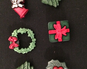 May include: A collection of Christmas-themed decorative buttons. The set includes a green Christmas tree, a green wreath with a red bow, red bells with a silver bow, a green holly leaf with red berries, and a green gift box with a red bow.