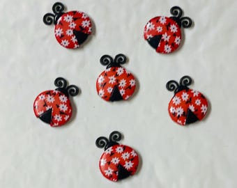 May include: Six red ladybug-shaped embellishments with white floral patterns and black accents. Each ladybug has a black head and antennae. The ladybugs are arranged on a white surface.