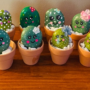 May include: Nine painted rock cactus decorations in small terracotta pots. Each rock is painted green with white dots and a cute face. Some have additional details like flowers or hearts.