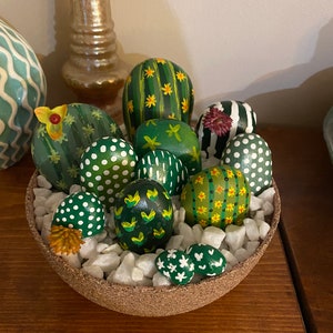 May include: A collection of painted rocks arranged to look like a cactus plant. The rocks are painted in shades of green, yellow, and white with various patterns. The rocks are sitting in a small brown bowl filled with white pebbles.