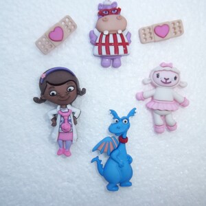 May include: A collection of colorful character figurines, including a purple hippo, a lamb, a blue dragon, and a girl in a doctor's coat. Two bandage-shaped embellishments with pink hearts are also present. These are likely decorative items.