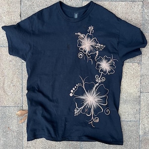 May include: A black short-sleeve t-shirt featuring a bleached design of three hibiscus flowers arranged vertically on the right side. The floral pattern is created with light beige, faded lines against the dark fabric, showcasing detailed petals.