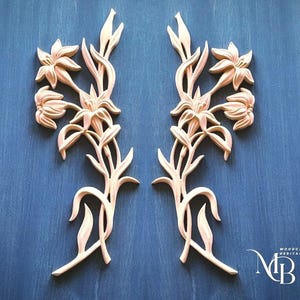 May include: Two symmetrical light-colored wooden carvings featuring detailed lily flower motifs with stems and leaves. The carvings are arranged vertically, mirroring each other. A small logo with the text Woodcarving Heritage MB is in the corner.