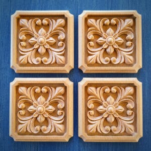 Puede incluir: Cuatro baldosas cuadradas de madera clara con un motivo floral tallado. Cada baldosa muestra una flor central rodeada de patrones de hojas simétricas en espiral dentro de un marco de borde elevado, dispuestas en una cuadrícula de dos por dos.