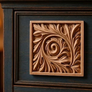 May include: A square wooden applique featuring a detailed acanthus leaf carving with a central spiral design. The natural light-colored wood piece is mounted onto a dark blue painted wooden surface with a visible grain and distressed finish.