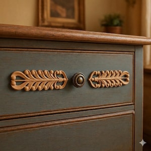 May include: A blue-grey wooden drawer front featuring a natural wood-toned top edge and two horizontal, symmetrical, carved wooden leaf-shaped decorative appliques flanking a single, round, antique brass-finished metal knob in the center.