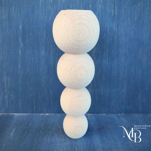May include: A wooden, unfinished decorative column composed of four stacked spheres. The top sphere has a hollowed-out center. The column is set against a blue background. The bottom right corner has a logo that says "Woodcarving Heritage" and the initials "NB".