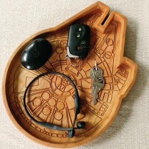 May include: A wooden tray in the shape of the Millennium Falcon, with etched details. The tray holds a black wireless earbud, a car key fob, a set of keys, and a black pebble-shaped object.