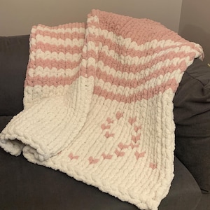 May include: A soft, chunky knit blanket in white and light pink. The blanket features horizontal stripes of pink and white, with a pattern of pink hearts on the lower portion. The blanket is folded over a gray surface.