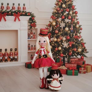 May include: Two handmade crochet dolls dressed in red and white marching band outfits stand in front of a decorated Christmas tree. The dolls have blonde and dark hair, and wear matching hats and boots. A smaller doll is also present.
