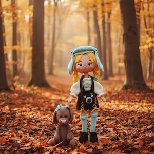 May include: A crocheted doll and dog are set in a forest scene with autumn leaves. The doll has blonde hair, a blue hat, and a camera-themed outfit. The dog is brown with a blue bow. The background features tall trees and sunlight.