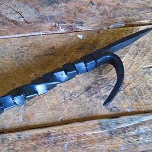 May include: A black, hand-forged metal fire poker with a twisted shaft and a hooked end. The tool is designed for managing embers and logs in a fireplace or fire pit. The poker rests on a weathered wooden surface.