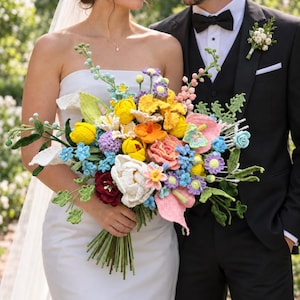 May include: A large, colorful bridal bouquet made of crocheted flowers in shades of yellow, pink, blue, white, and deep red. The arrangement features various textured blooms, green crocheted leaves, and stems, held together in a bundle.