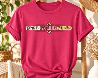 May include: A bright pink short-sleeved t-shirt featuring the text "UNBOTHERED. UNFILTERED. OVERHEATED." printed in a stylized font with a decorative floral-inspired graphic element above and below the words in shades of yellow, green, and pink.
