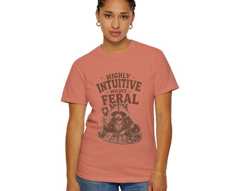 May include: A coral-colored short-sleeve t-shirt featuring a dark brown graphic of a raccoon dressed in robes, holding a tarot card, and sitting before a crystal ball. The text above the raccoon reads HIGHLY INTUITIVE MILDLY FERAL.