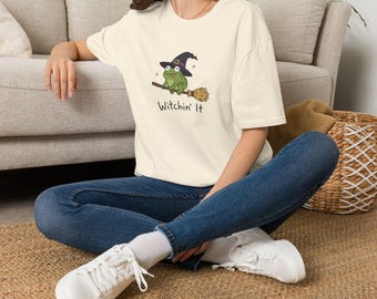 May include: An off-white short-sleeve t-shirt featuring a graphic of a green frog wearing a purple witch hat and riding a brown broomstick. Below the frog, the text reads "Witchin' It" in a simple black font. The shirt is paired with blue jeans.