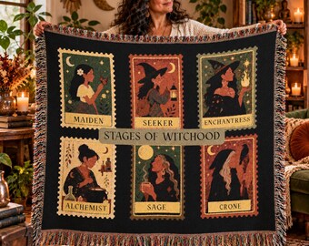 May include: A black woven throw blanket featuring six rectangular panels with illustrations of figures labeled Maiden, Seeker, Enchantress, Alchemist, Sage, and Crone. The center text reads STAGES OF WITCHOOD. The blanket has a fringed edge.