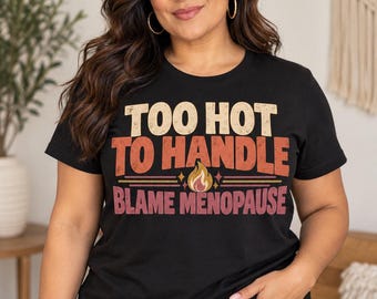 May include: A black short-sleeved t-shirt featuring the text "TOO HOT TO HANDLE" in beige, "BLAME MENOPAUSE" in pink, and a small central flame graphic. The design uses a distressed, vintage-inspired font style on the front of the shirt.