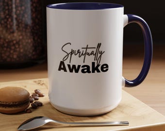 May include: White ceramic mug with a navy blue interior and handle. The mug features the words "Spiritually Awake" in black script. A spoon, coffee beans, and a pastry are on a wooden surface.
