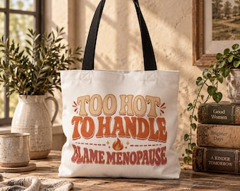 May include: A white canvas tote bag with black shoulder straps featuring the text "TOO HOT TO HANDLE BLAME MENOPAUSE" in a retro-style orange and red font, accompanied by a small flame graphic centered between the two lines of text.