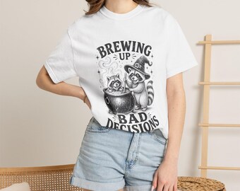 May include: A white short-sleeved t-shirt featuring a black and white graphic print of two raccoons, one wearing a witch hat, stirring a cauldron. The text above and below the illustration reads, "BREWING UP BAD DECISIONS" in a bold font.