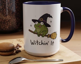 May include: A white ceramic mug with a dark blue interior and handle. The front features a cartoon green frog wearing a black witch hat, riding a brown broomstick. Below the illustration, the text reads Witchin' It in black lettering.